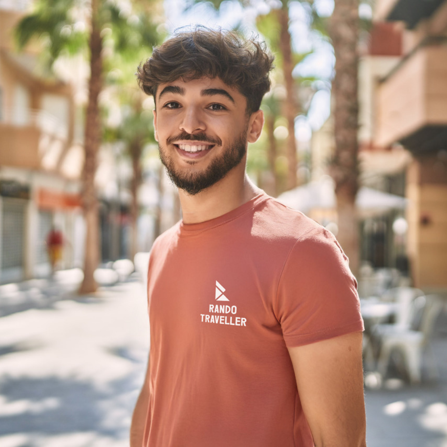 Man wearing a 'Rando Traveller' t-shirt standing outdoors with palm trees and buildings in the background.